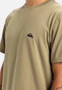 Camiseta de algodón verde oliva con cuello redondo y mangas cortas, con un pequeño logotipo negro bordado en el pecho.