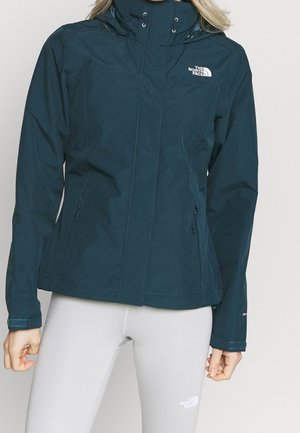 Kobieta w ciemnoniebieskiej kurtce z kapturem North Face i jasnoszarych legginsach North Face na jednolitym tle.