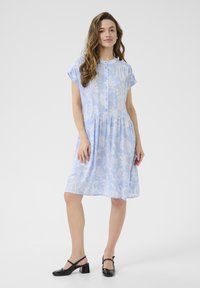 Jasnoniebieska sukienka w wzór paisley, krótki rękaw, zapinana na guziki z gromadzonym wcięciem w talii. Noszona z czarnymi butami na obcasie typu slingback.