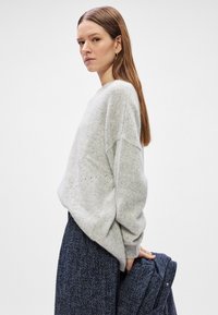 Suéter de malha cinza claro com um corte solto e ombros descaídos. O tecido tem um aspeto suave e texturizado. Combinado com uma saia estampada azul escura.