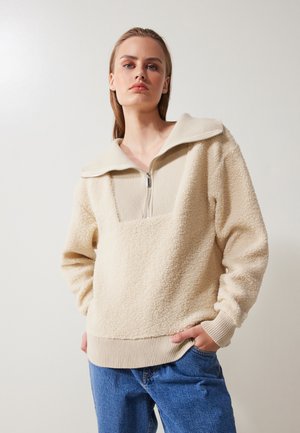 Cream-colored fleece pullover with a ribbed collar and zip closure, featuring a textured surface and ribbed cuffs, paired with blue jeans.