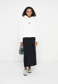 Haut à capuche blanc avec logo noir, jupe noire superposée avec poches, sac à main vert et chaussures à motifs gris clair. Design simple et moderne.