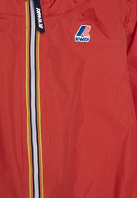 Raudona lengva striukė su užtrauktuku priekyje, su daugiaspalve juostele ir K-Way logotipu viršuje kairėje. Sklandi tekstūra, laisvalaikio dizainas.