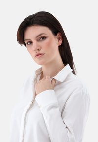 Weißbluse mit gefransten Kanten, glatter Textur und einem einzelnen goldenen Knopf am Bündchen. Die Hand des Models hält sanft den Kragen.