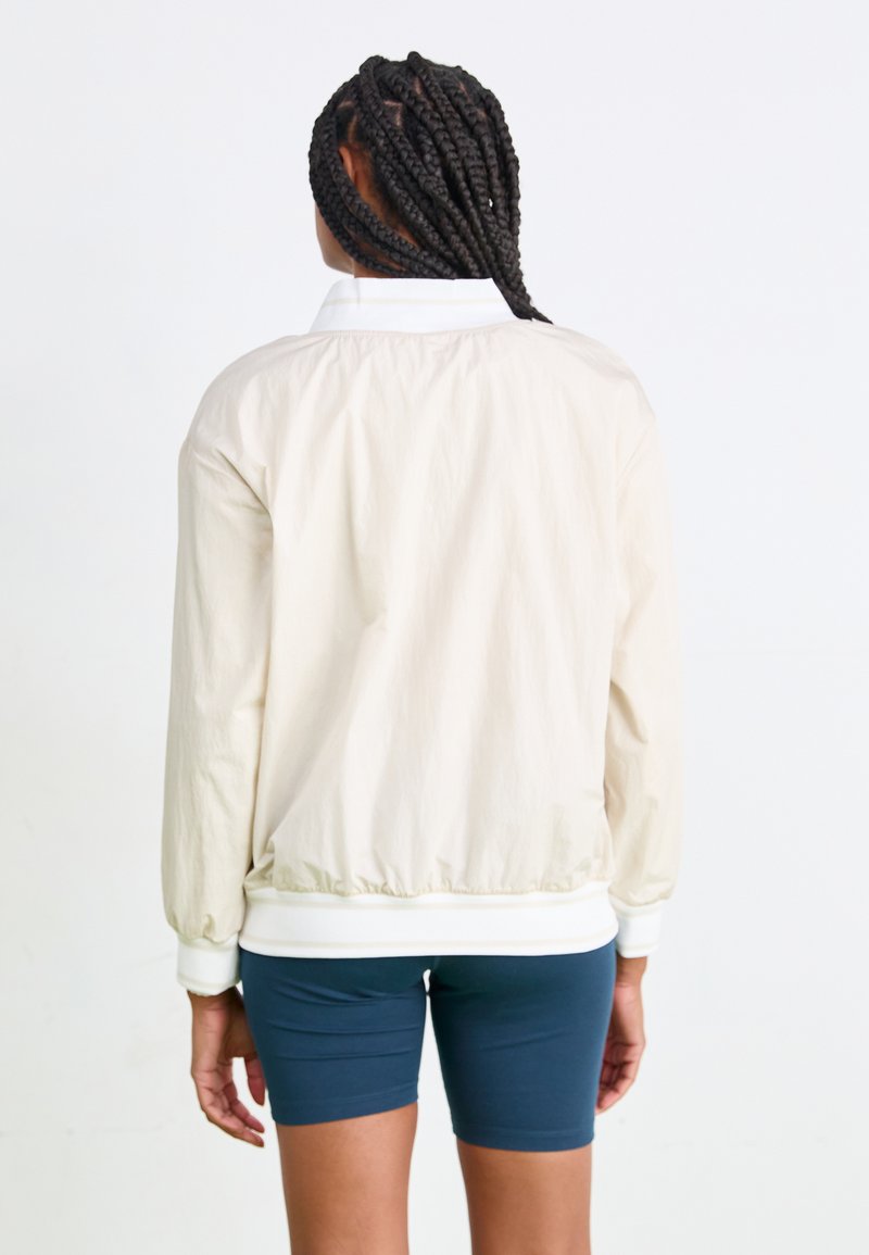 Jasno beżowa kurtka bomberka z marszczonym dołem i mankietami, z białym kołnierzykiem. Prezentowana z tyłu na granatowych szortach.