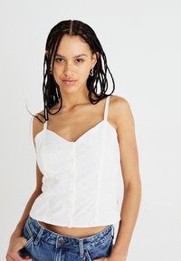 Bijeli pamučni crop top s uzorkom prozora, s prednjim dugmadi i tankim trakama, uparen s trapericama.
