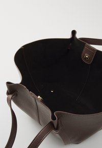 Sac fourre-tout en cuir marron avec un extérieur texturé, une ouverture en haut et un intérieur en daim. Fermeture avec un bouton-pression doré et des anses en cuir. Design minimaliste.