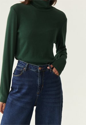 Personne portant un pull à col roulé côtelé vert foncé et un jean taille haute en denim bleu avec une main dans la poche.