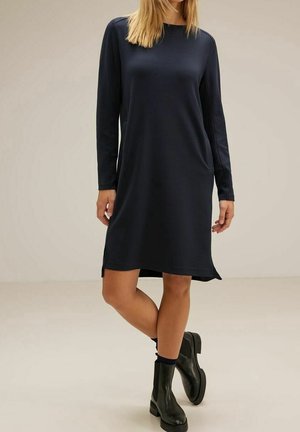 Robe longue bleu marine à manches longues en tissu doux, avec une coupe décontractée, un col rond et des fentes latérales, portée avec des bottines noires.