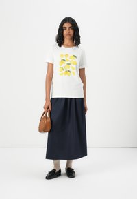 Weißes T-Shirt mit Zitronenmotiv, kurze Ärmel; kombiniert mit einem marineblauen knöchellangen Rock und einer braunen Handtasche; schwarze Schuhe und beigefarbene Socken.