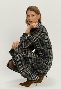 Žena v kľaku, oblečená v dlhých kšaňových šatách s pruhmi a hnedými semišovými vysokými podpätkami, pozerajúca sa na kameru s neutrálnym pozadím.