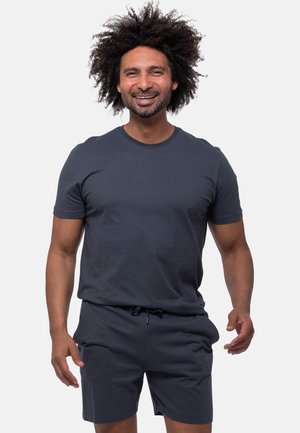 Lächelnder Mann mit lockigem Haar trägt ein dunkelgraues T-Shirt und passende Shorts und steht vor einem einfarbigen hellen Hintergrund.