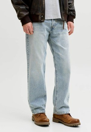 Muž nosi svetlomodré rovné rifle, hnedé kožené topánky, biele tričko a tmavohnedú koženú bundu, stojí na bielom pozadí.