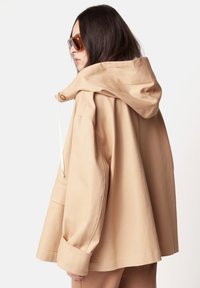 Beige Kapuzenjacke mit lässigem Schnitt, großen Taschen und einem Kordelzugdetail an der Kapuze. Glatte Stoffqualität und weite Ärmel.