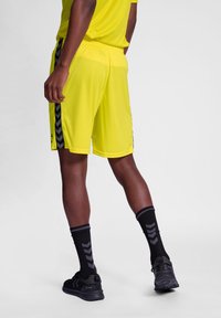 Gelbe Sportshorts mit schwarzen Seitenelementen und einer strukturierten Oberfläche, kombiniert mit schwarzen Socken im Chevron-Muster und schwarzen Laufschuhen.