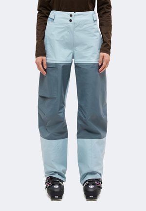 Un mannequin porte un pantalon de ski bicolore bleu clair et gris ardoise, avec des poches cargo et des chaussures de ski noires, sur un fond uni.