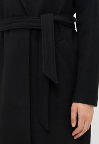 Cappotto di lana nera con cintura annodata e tasca laterale, con braccio e mano destra visibili su uno sfondo neutro.