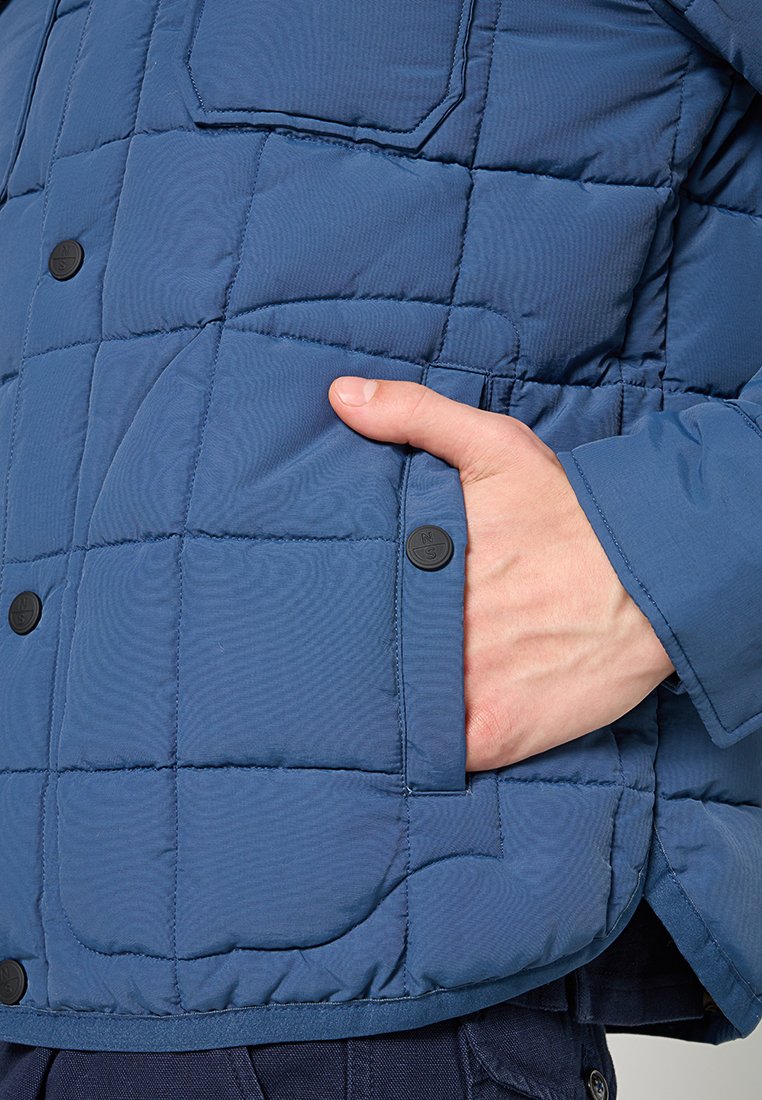Giacca trapuntata blu in stile puffer con una mano in una tasca laterale. Presenta bottoni a pressione neri e una texture liscia e lucida.
