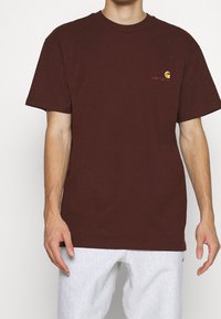 Burgundin värinen puuvillapaita, jossa on pyöreä kaula-aukko, lyhyet hihat ja pieni keltainen ja punainen logo rinnassa. Sileä pinta.