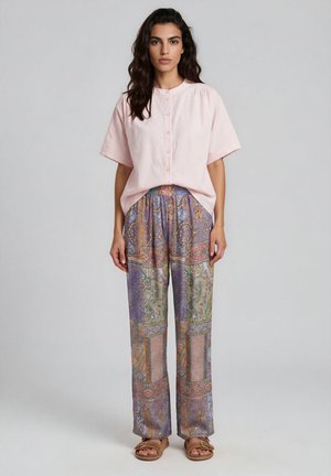 Woman wearing a light pink short-sleeve buttoned shirt, multicolored patterned loose pants, and brown sandals standing against a plain background.
