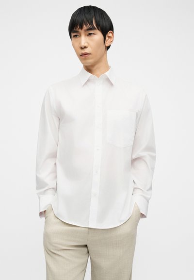 Ασιάτης άνδρας που φοράει λευκό πουκάμισο με κουμπιά και μακριά μανίκια και ανοιχτό μπεζ παντελόνι, στέκεται με τα χέρια στις τσέπες μπροστά σε απλό φόντο.