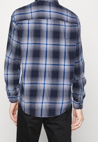 Camicia a maniche lunghe a quadri con motivi blu navy, bianchi e blu. Il tessuto ha una texture morbida, con polsini a bottone e orlo arrotondato.