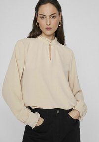 Beige långärmad blus med en rufflad krage och nyckelhålsdetalj. Har elastiska rufflade ärmslut och en mjuk, flödande tygstruktur.