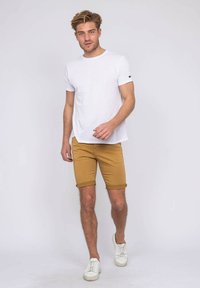 T-shirt blanc à manches courtes associé à un short jaune moutarde roulé et des baskets blanches. Le t-shirt présente un petit logo sur la manche.