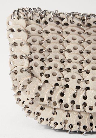 Krēma krāsas ādas soma ar apaļiem izgriezumiem, kas savienoti ar sudraba metāla gredzeniem, strukturēta dizaina ar slāņainu tekstūru, minimālistiska estētika.