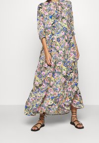 Robe maxi florale dans des nuances de vert, rose, bleu et jaune. Dotée de manches longues, d'un col boutonné et d'un ourlet en étages. Sandales noires à brides.