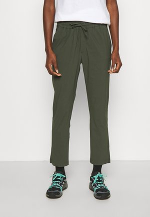 Personne portant un pantalon vert olive à cordon, des chaussettes noires et des chaussures de sport noires avec des lacets turquoise, debout sur un fond blanc.