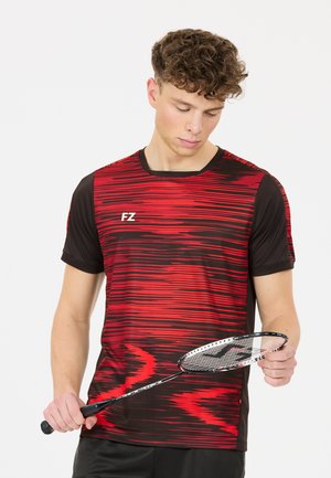 Nuori mies mustassa ja punaisessa urheilupaidassa pitää ja tutkii sulkapallomailaa yksiväristä valkoista taustaa vasten.