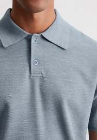 Jasnoniebieska koszulka polo wykonana z fakturowanej tkaniny, z kołnierzykiem, trzema guzikami i subtelnymi detalami ściegów. Zbliżenie na górną część.