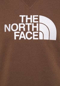 Sudadera marrón con diseño de escote en pico que presenta un gran logo blanco de "The North Face" y líneas de acento curvas. Textura suave.