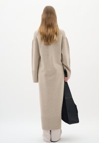 Lång beige stickad cardigan med ribbade muddar och nederkant, ihop med en svart axelväska. Mjuk textur, enkel design och neutrala färger.