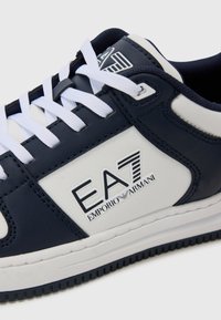 Marineblå og hvite sneakers med glatt lær øvre, med hvite lisser, logo detaljer og gummisåle. Minimalistisk design, lav profil.
