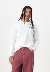 White long-sleeve t-shirt with a black logo on the chest, paired with pink trousers. The fabric appears smooth with a relaxed fit.