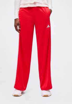 Person iført lyse røde Adidas træningsbukser og hvide sneakers står med hænderne i lommerne på en ensfarvet hvid baggrund.