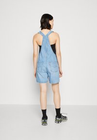 Pettorina in denim blu con vestibilità comoda, spalline regolabili e due tasche posteriori, abbinata a una maglietta nera e scarpe da ginnastica nere.