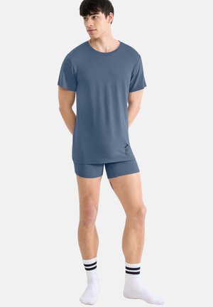 Homme portant une chemise à manches courtes bleu ardoise et un short assorti, chaussettes blanches avec des rayures noires, debout devant un fond clair uni.