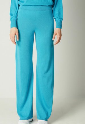 Femme portant un pantalon en maille turquoise ample et des baskets blanches, debout devant un fond clair uni.