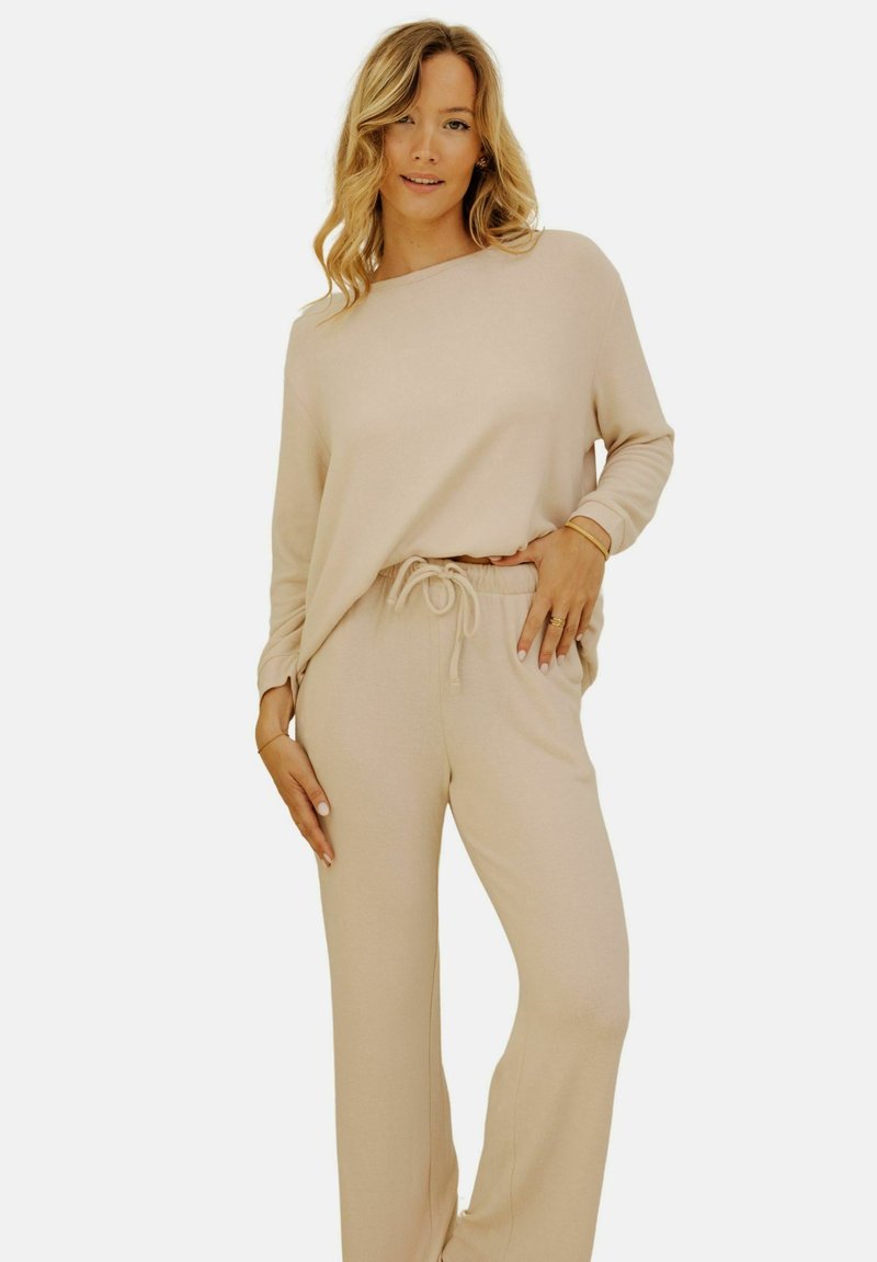 Femme posant, portant un haut beige à manches longues et un pantalon à cordon assorti, avec des cheveux blonds ondulés et des bijoux minimalistes sur fond blanc.