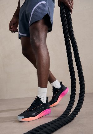 Zapatilla deportiva negra con acentos en rosa y gris, parte superior texturizada y suela de goma. El modelo lleva pantalones cortos grises y calcetines blancos, sosteniendo una cuerda negra.