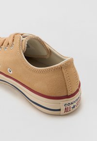 Zapatilla de ante marrón con forro blanco, rayas rojas y azules en la suela, ojales de metal y logo "Converse All Star" en el talón.