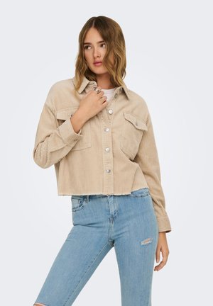 Ung kvinne med bølget hår iført en beige kort kordfløyelsjakke og lys blå revne skinny jeans mot en ensfarget bakgrunn.