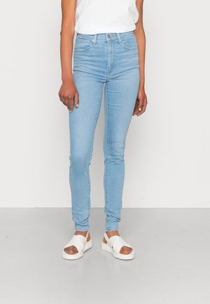 Personne portant un jean skinny bleu clair, des sandales blanches avec des larges brides, et un top blanc sans manches rentré, debout sur un sol gris.