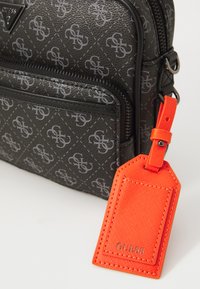 Black Guess bag with repeating logo pattern, front zipper pocket, and attached bright orange luggage tag with Guess branding.