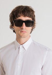 Occhiali da sole neri rettangolari con una montatura spessa, indossati sopra una camicia bianca a maniche lunghe. La camicia presenta un colletto classico e bottoni.