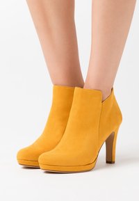 Keltaiset mokkanahkasaappaat nilkkapituisena, joissa on sileä pinta, pyöreät varpaat ja korkea stiletto-korko. Saappaissa on kaareva kaulus ja vähäiset ompeluyksityiskohdat.