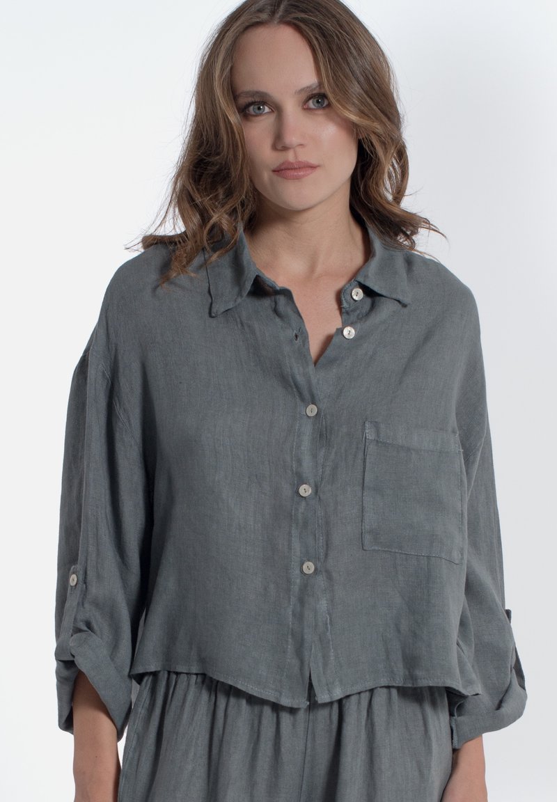Camicia in lino grigia con vestibilità rilassata, chiusura a bottoni sul davanti, colletto a punta, taschino sul petto e maniche arrotolate. Texture morbida e design leggero.